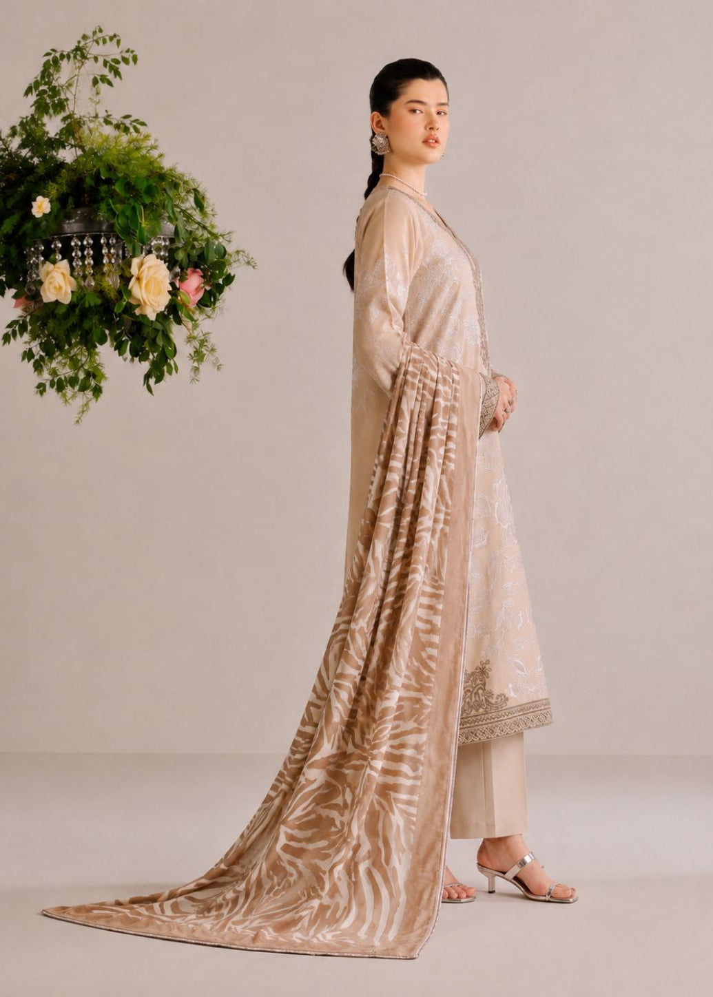 A woman models a traditional beige and gold embroidered suit with a long patterned dupatta, standing against a neutral studio backdrop next to a hanging floral arrangement.
