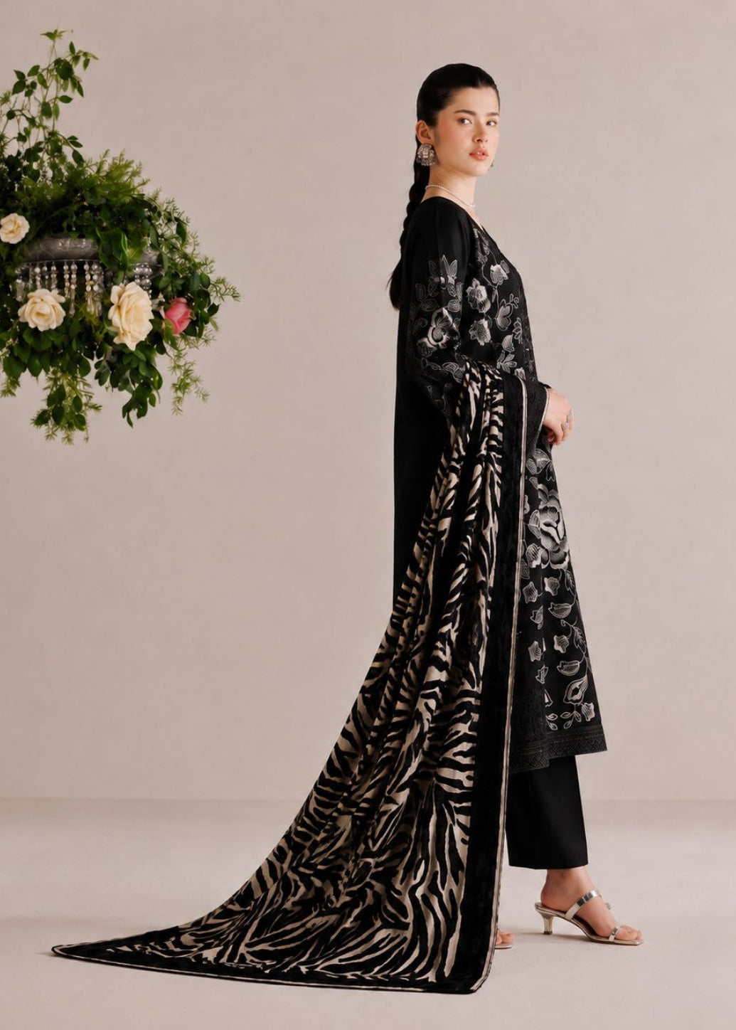 A woman models a black three-piece salwar kameez suit with intricate silver floral embroidery and a matching black-and-silver patterned dupatta, set against a neutral beige background with a hanging floral arrangement.