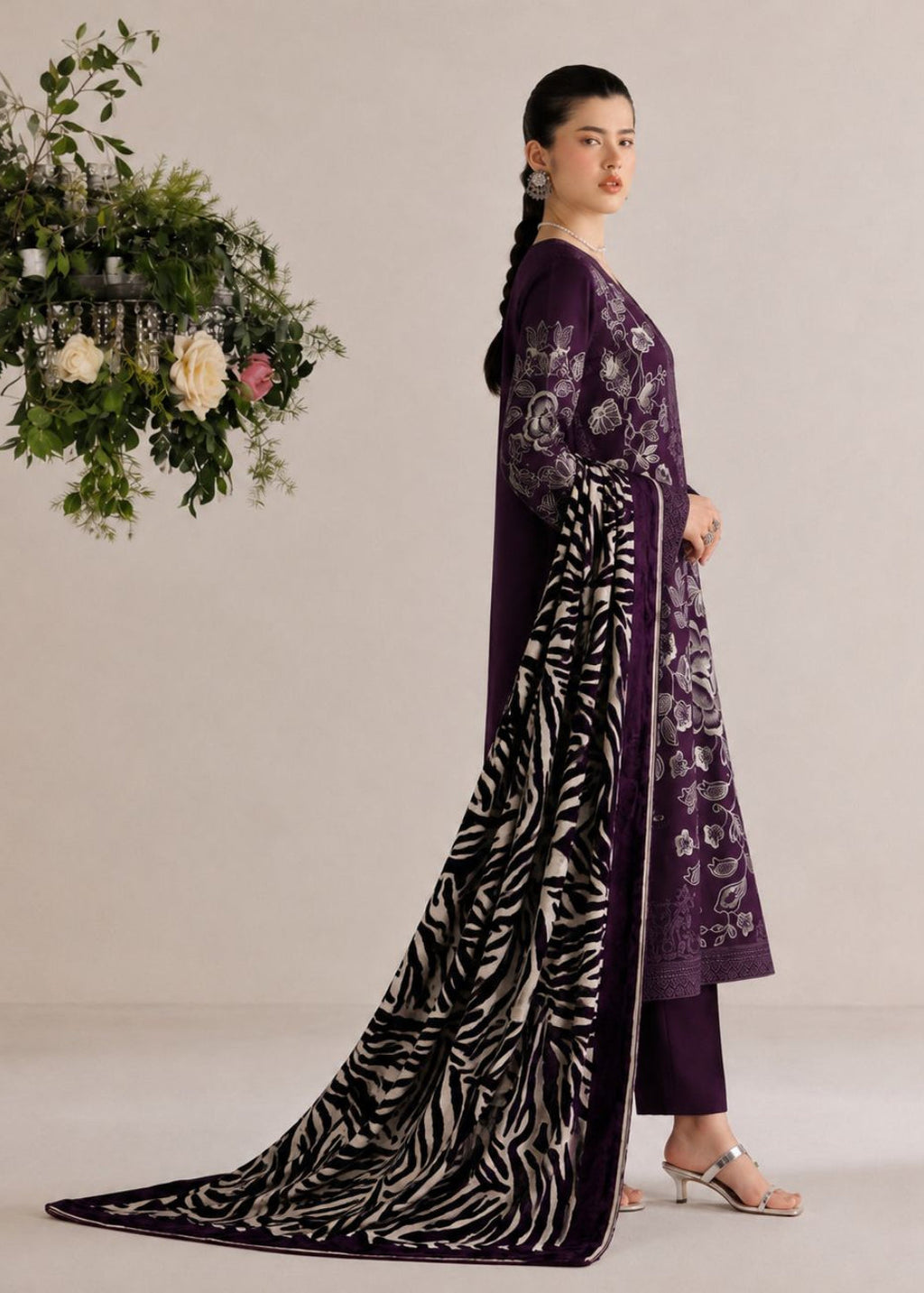 A woman models a traditional Indian outfit consisting of a deep purple kurta with floral embroidery, matching purple trousers, and a long black dupatta with white zebra-like stripes, posed against a neutral beige background with a hanging floral arrangement.