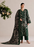 A model stands in a studio wearing a dark green traditional outfit with floral embroidery, matching pants, and a long patterned dupatta draped over the floor. A hanging floral arrangement is visible to the left against a plain light beige background.