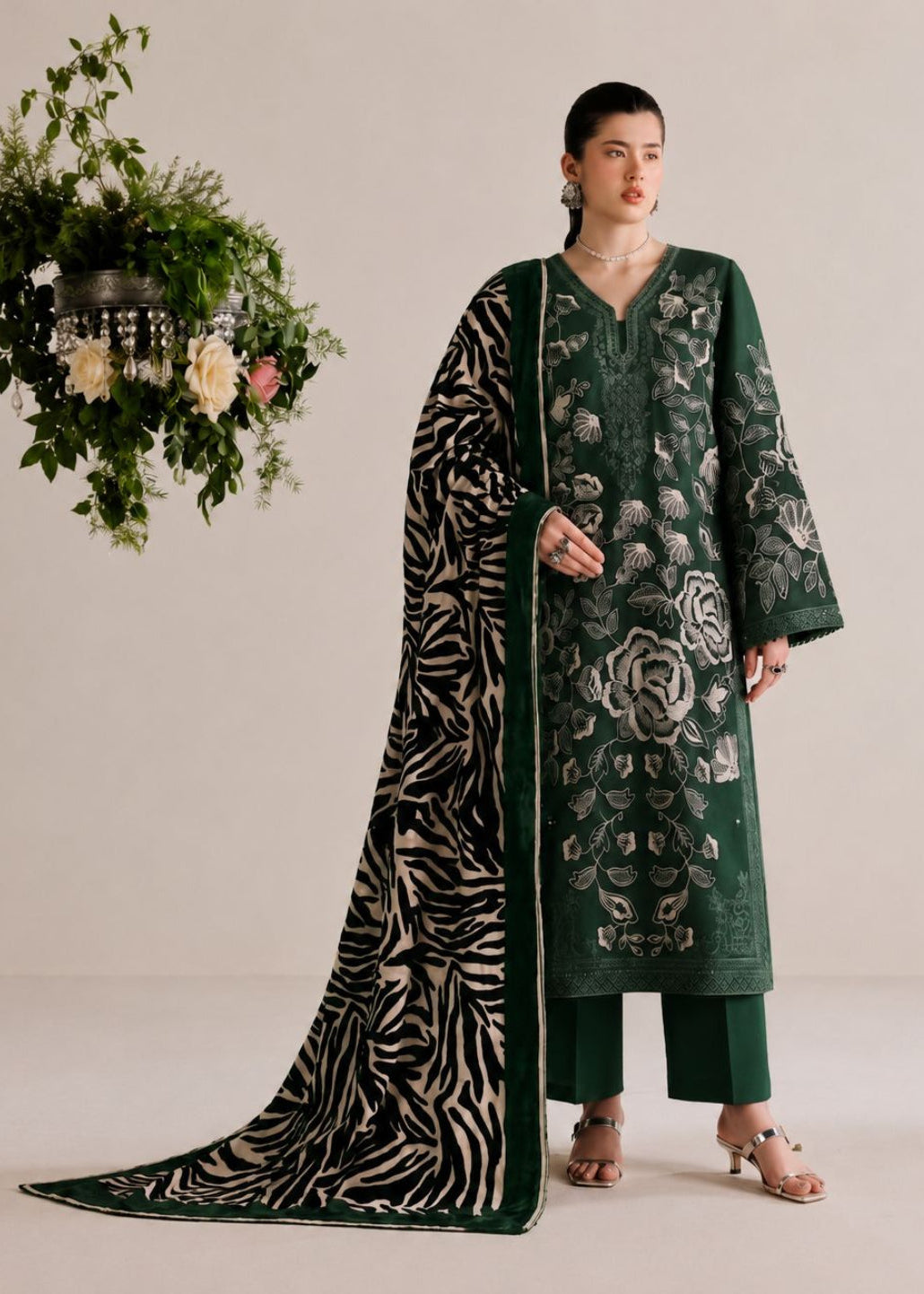 A model stands in a studio wearing a dark green traditional outfit with floral embroidery, matching pants, and a long patterned dupatta draped over the floor. A hanging floral arrangement is visible to the left against a plain light beige background.