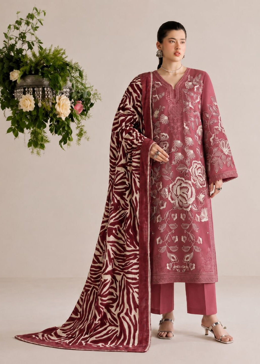 A woman models a traditional three-piece salwar kameez suit in dusty rose pink with beige floral embroidery, paired with matching trousers and a long dupatta featuring a contrasting beige zebra print on a deep pink background. She is posed against a neutral beige studio backdrop with a decorative hanging floral arrangement to the side.