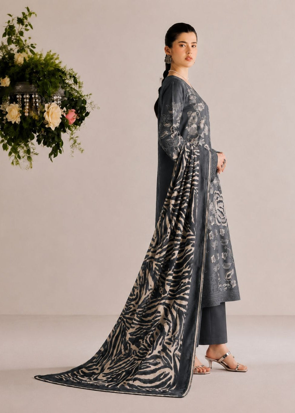 A woman models a dark gray three-piece traditional outfit with a zebra-print dupatta, standing against a neutral beige studio background with a decorative hanging floral arrangement on the left.