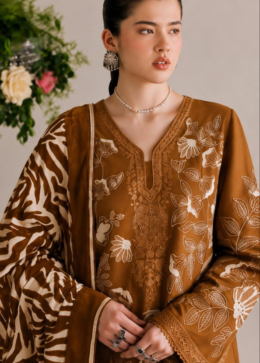 A woman wearing a brown and beige embroidered traditional outfit with a patterned dupatta, photographed against a soft neutral background with floral decor.