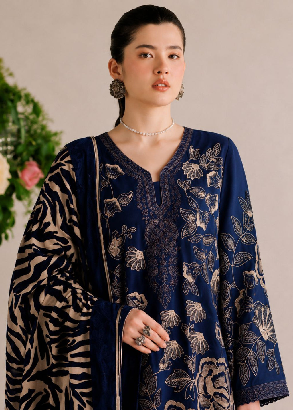 A woman models a navy blue embroidered kurta with beige floral patterns, paired with a matching patterned dupatta and jewelry, set against a neutral background with a floral arrangement.