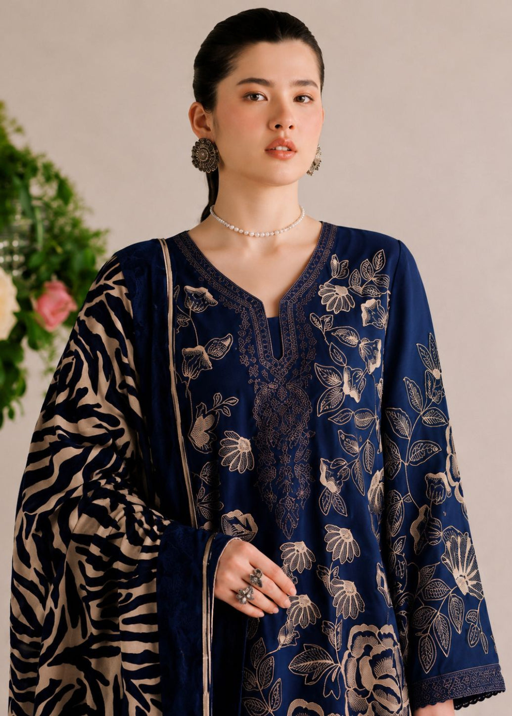 A woman models a navy blue embroidered kurta with beige floral patterns, paired with a matching patterned dupatta and jewelry, set against a neutral background with a floral arrangement.