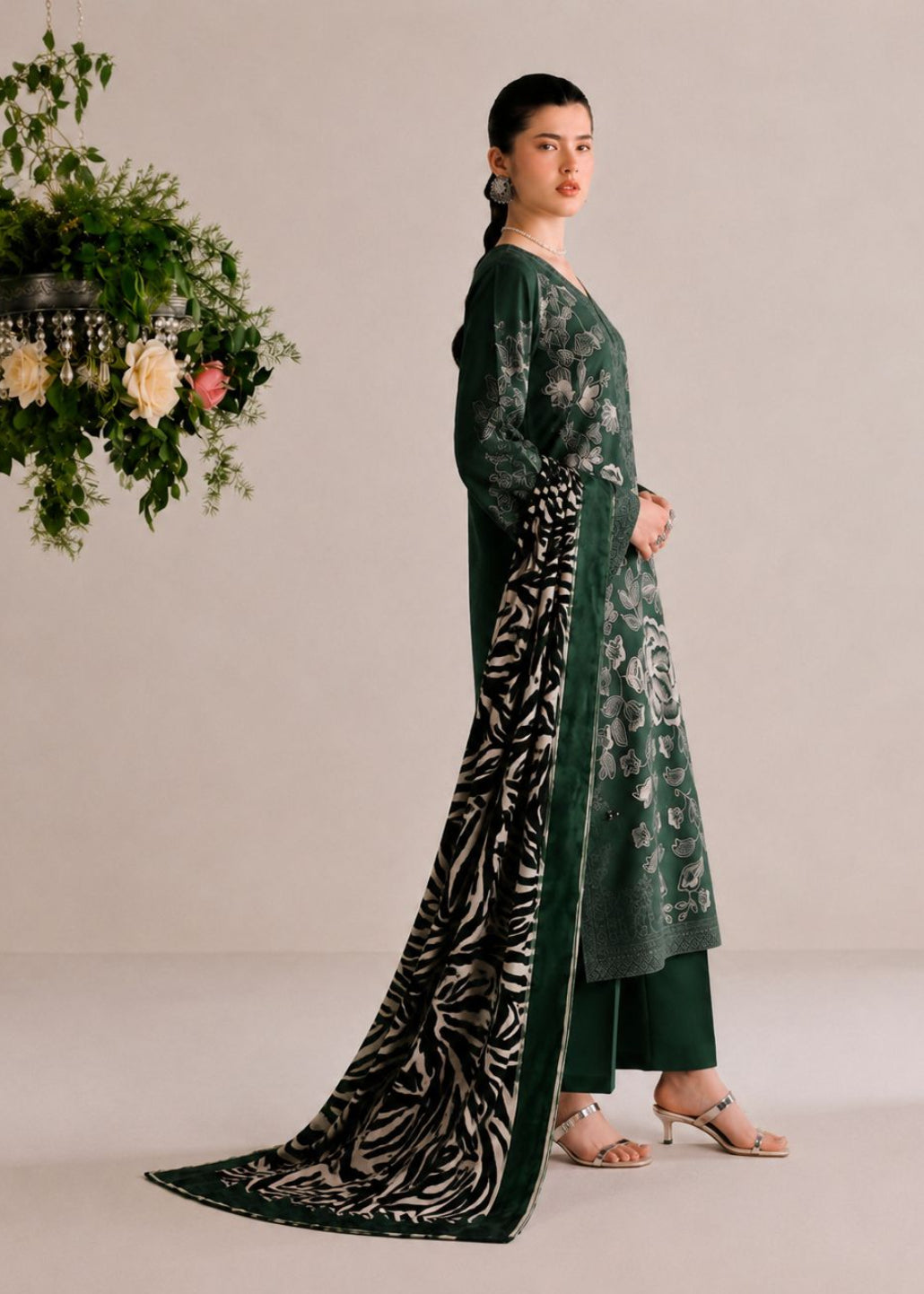 A woman models a dark green ethnic salwar kameez suit with floral prints and a black-and-white patterned dupatta, posed against a neutral beige studio backdrop with a hanging floral arrangement on the left.