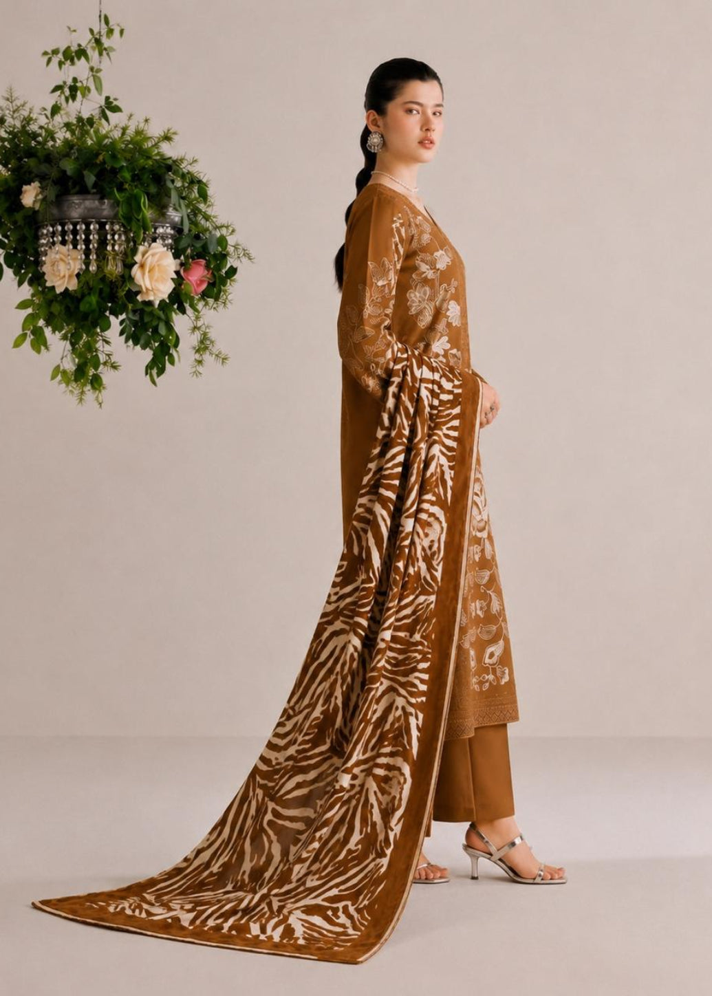 A woman models a three-piece brown ethnic suit with a matching printed dupatta, standing against a neutral beige studio backdrop with a hanging floral arrangement to the side.