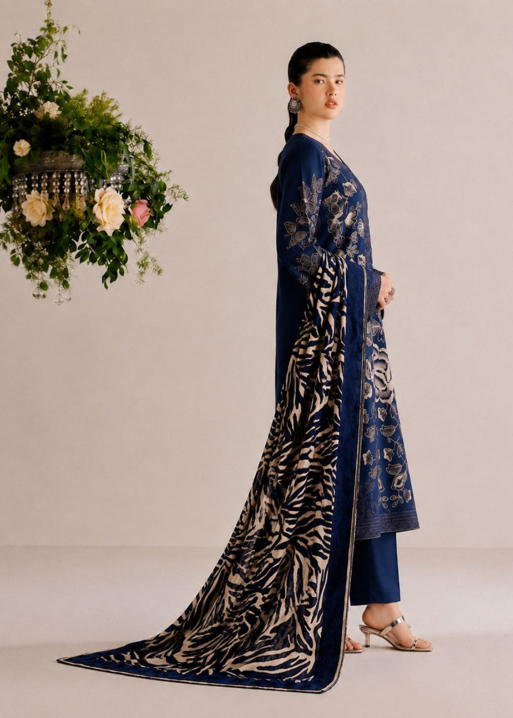 A woman models a navy blue traditional outfit with beige floral embroidery and a matching dupatta featuring a beige and navy abstract pattern, set against a neutral studio backdrop with a hanging floral arrangement.