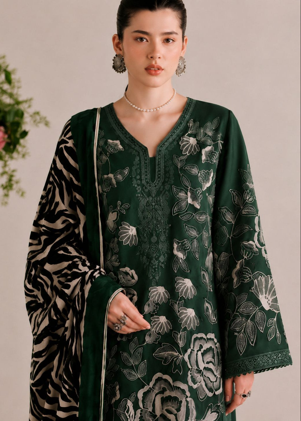 A woman modeling a dark green embroidered kurta with white floral patterns and a matching patterned dupatta, set against a neutral beige background with a floral prop on the left.