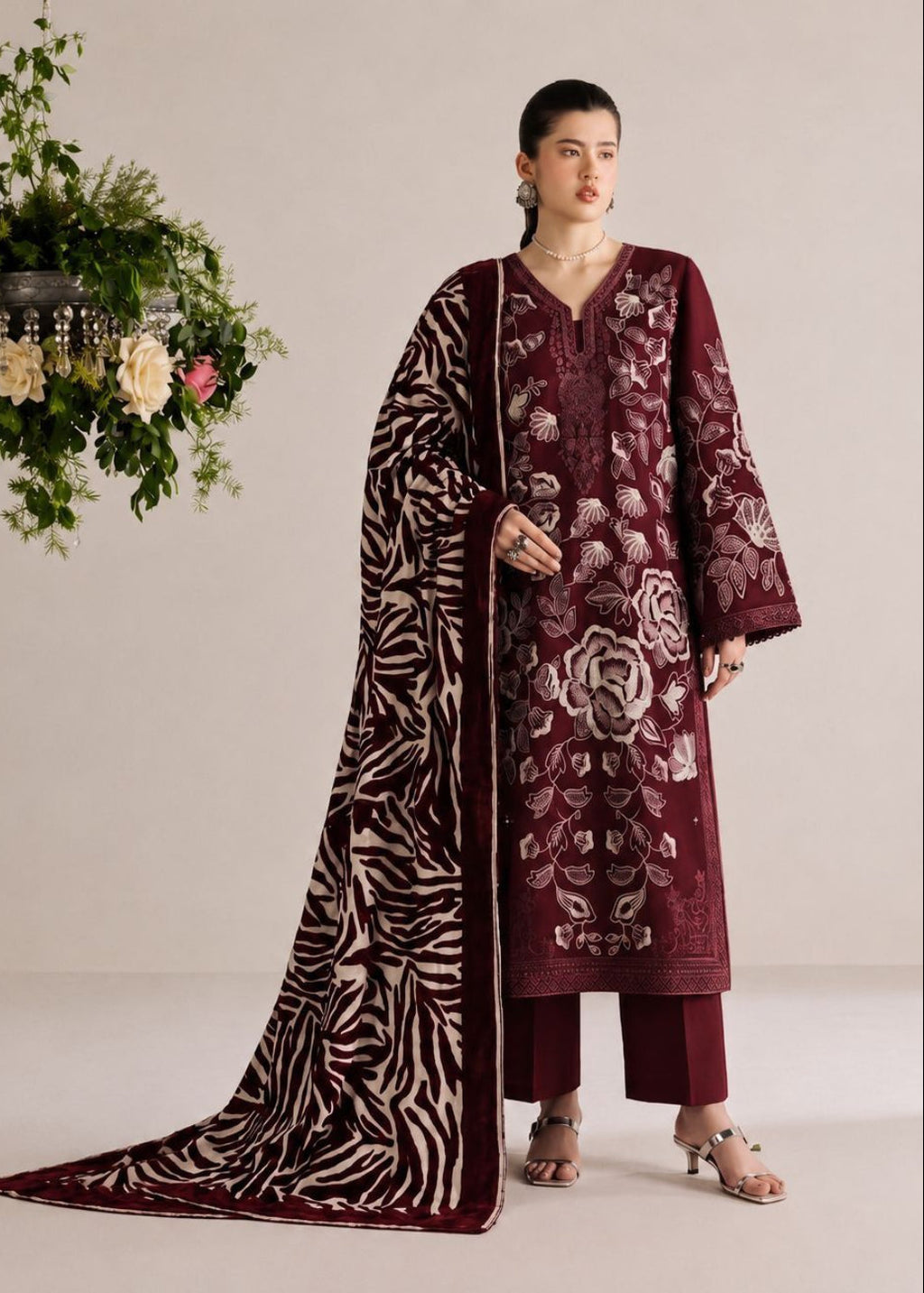 A woman models a traditional South Asian salwar kameez suit set in deep maroon, featuring floral embroidery on the kurta, matching trousers, and a long dupatta with a zebra print pattern, set against a neutral studio background with a hanging floral arrangement.