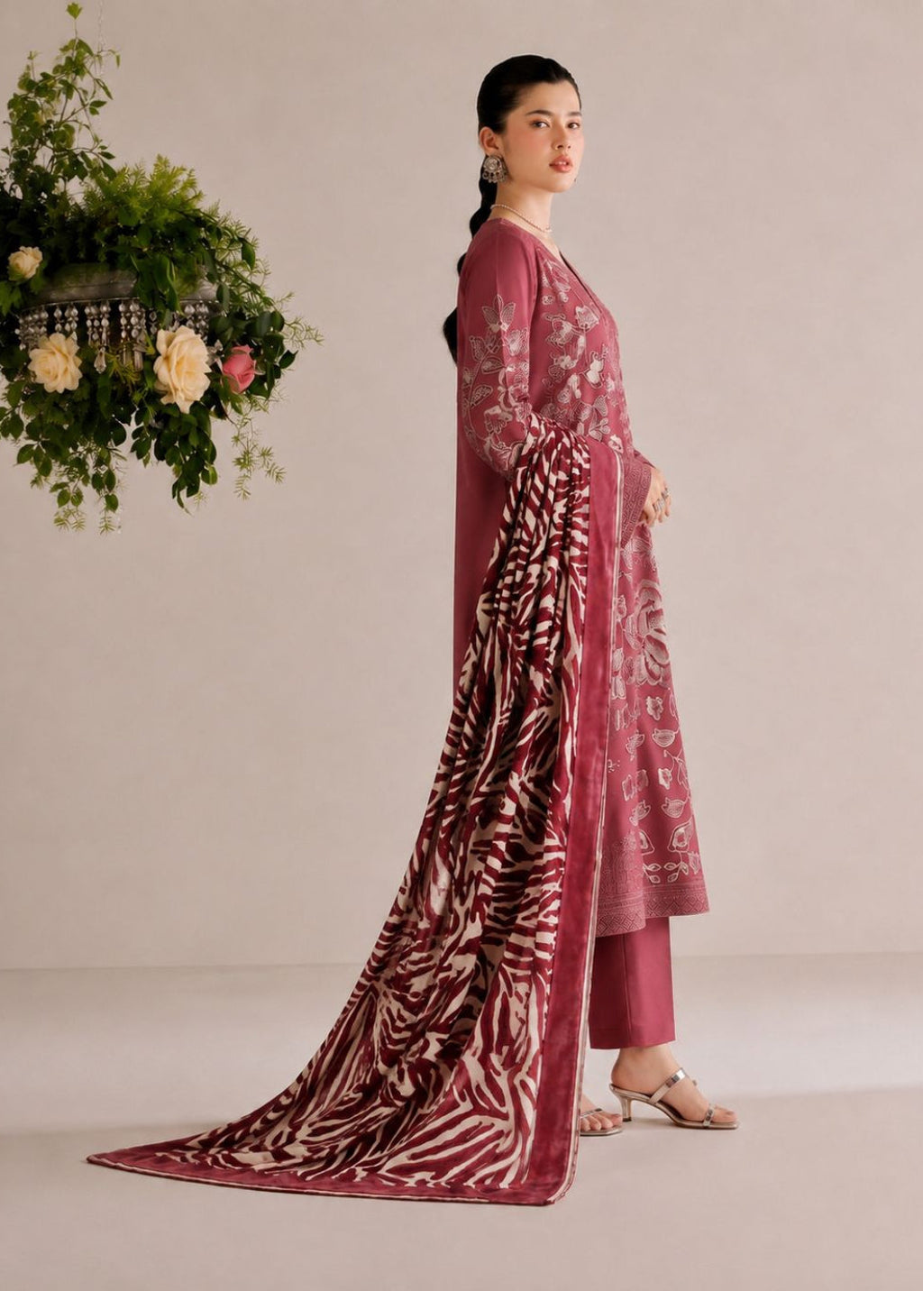 A woman models a traditional Indian salwar kameez suit in dusty rose pink with white floral embroidery, paired with matching trousers and a long dupatta featuring a white zebra print pattern, set against a neutral beige background with a hanging floral arrangement.