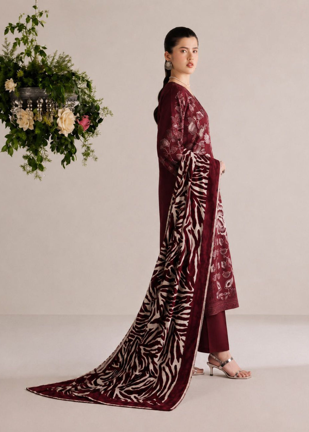 A woman models a traditional three-piece salwar kameez suit in deep maroon with intricate floral embroidery and a matching printed dupatta, posed against a neutral beige studio backdrop with a hanging floral arrangement.