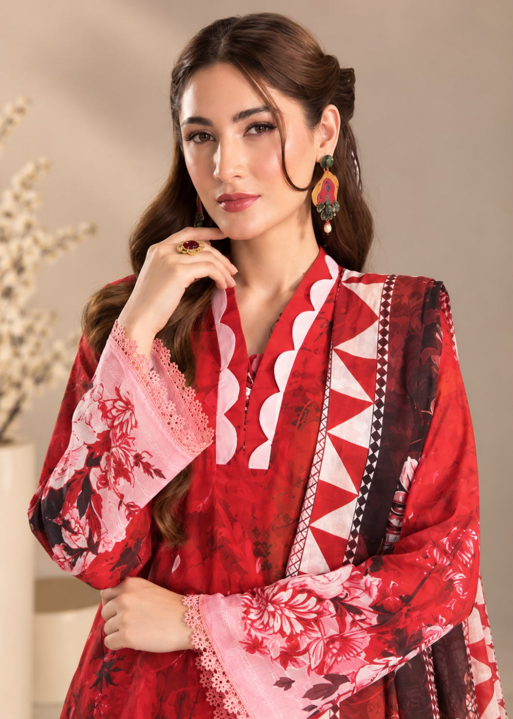 A woman modeling a red and pink printed traditional outfit with floral sleeves, a geometric-patterned border, and a matching dupatta against a neutral beige background.