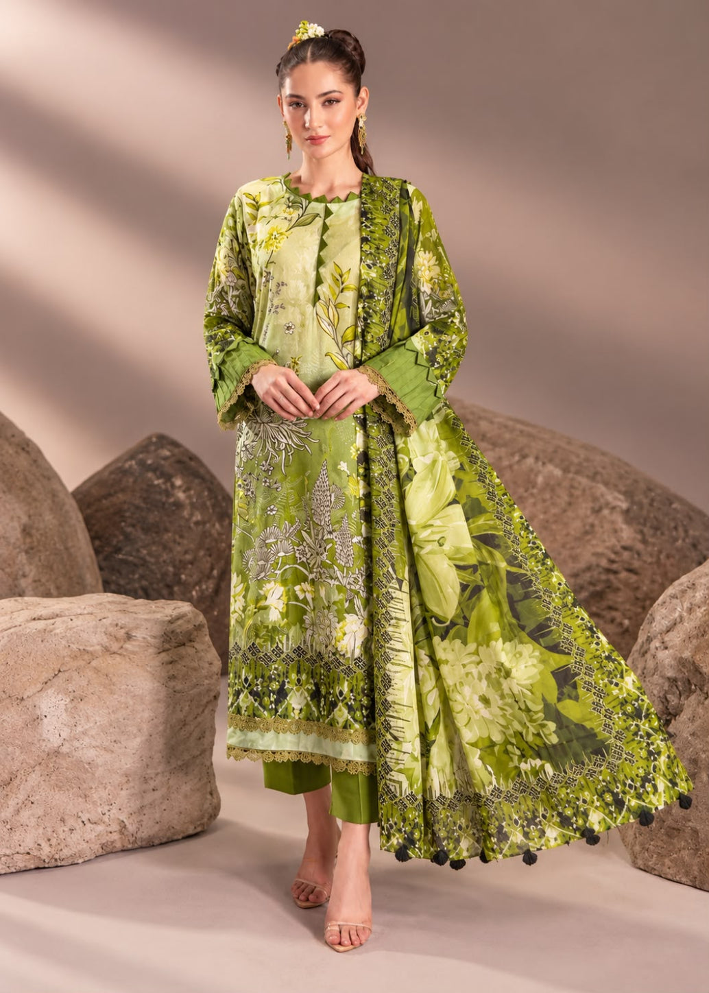 A woman models a three-piece green ethnic suit with floral embroidery and a matching dupatta, posed in front of large decorative rocks against a neutral beige backdrop.