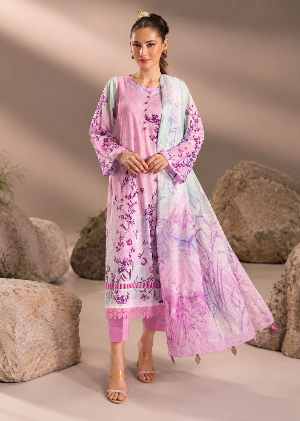 A woman models a traditional three-piece salwar kameez set in a studio setup with decorative rocks. The set features a light pink and lavender kurta with purple floral prints, a matching light blue-green dupatta with purple floral embroidery, and solid pink trousers.