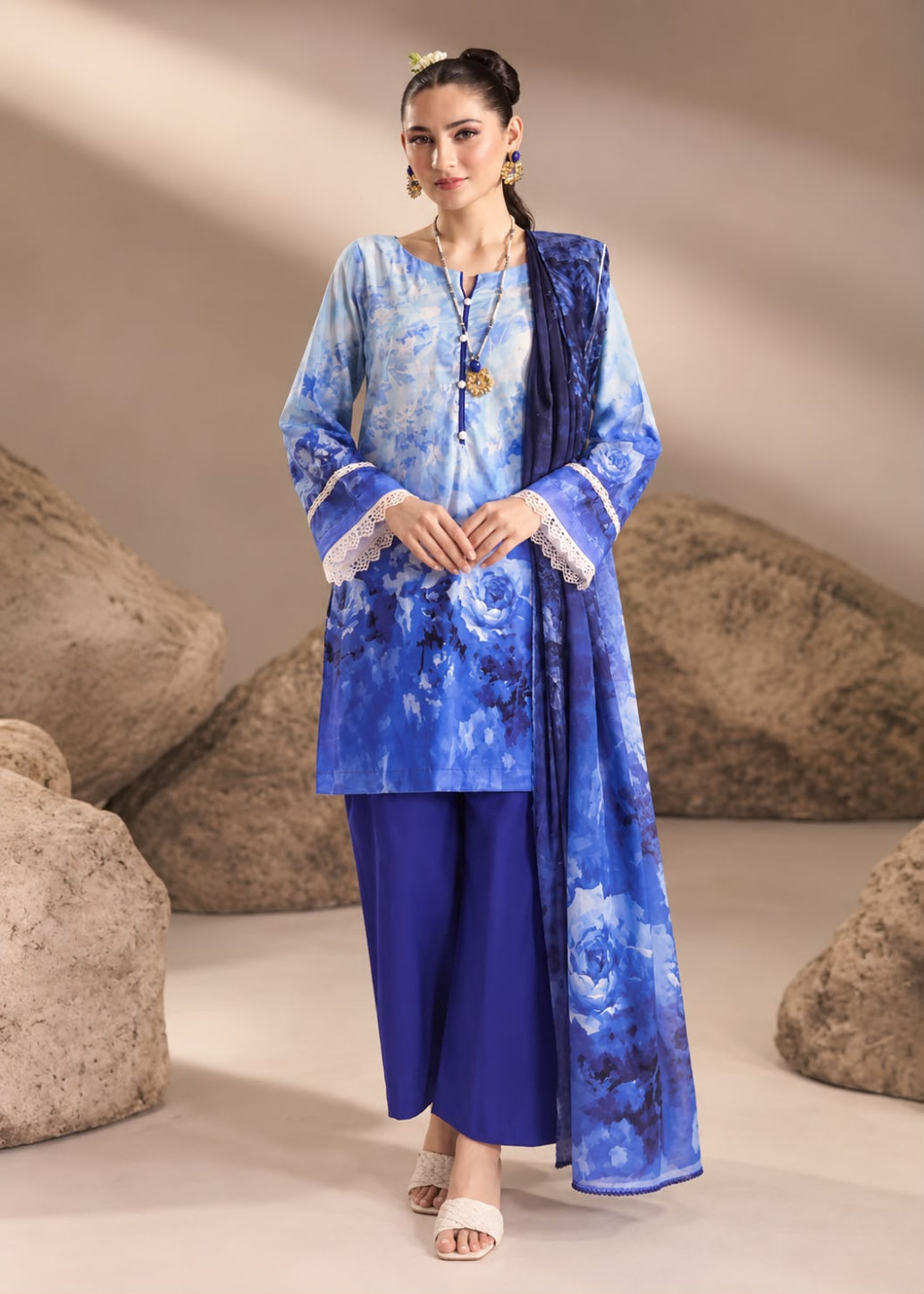 A woman models a traditional blue Indian outfit consisting of a blue floral printed kurta, solid royal blue pants, and a matching blue dupatta, set against a studio backdrop with large decorative rocks.