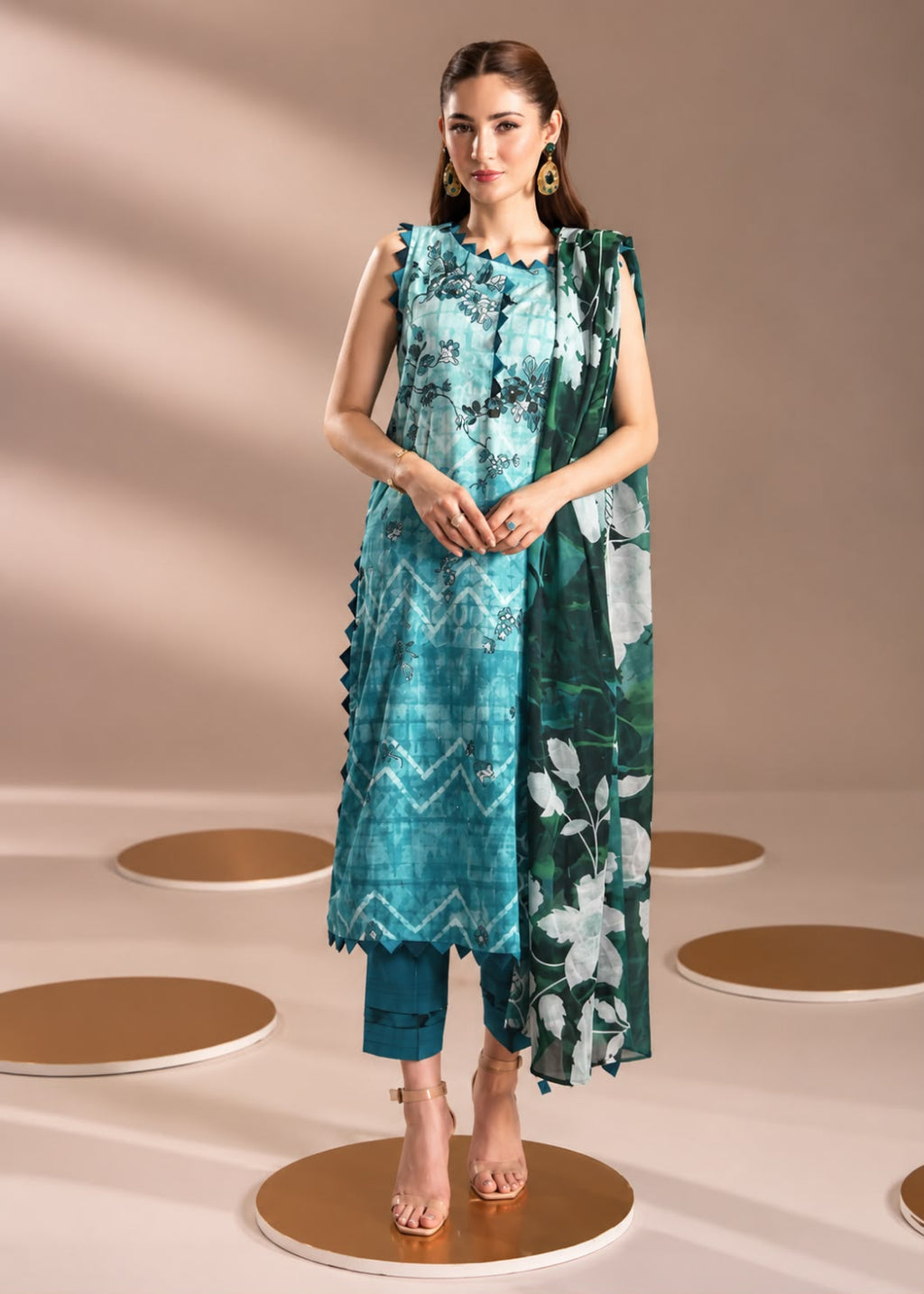 A woman models a three-piece teal and green ethnic suit set, featuring a sleeveless printed kurta, matching pants, and a floral dupatta, standing on circular gold platforms against a beige background.