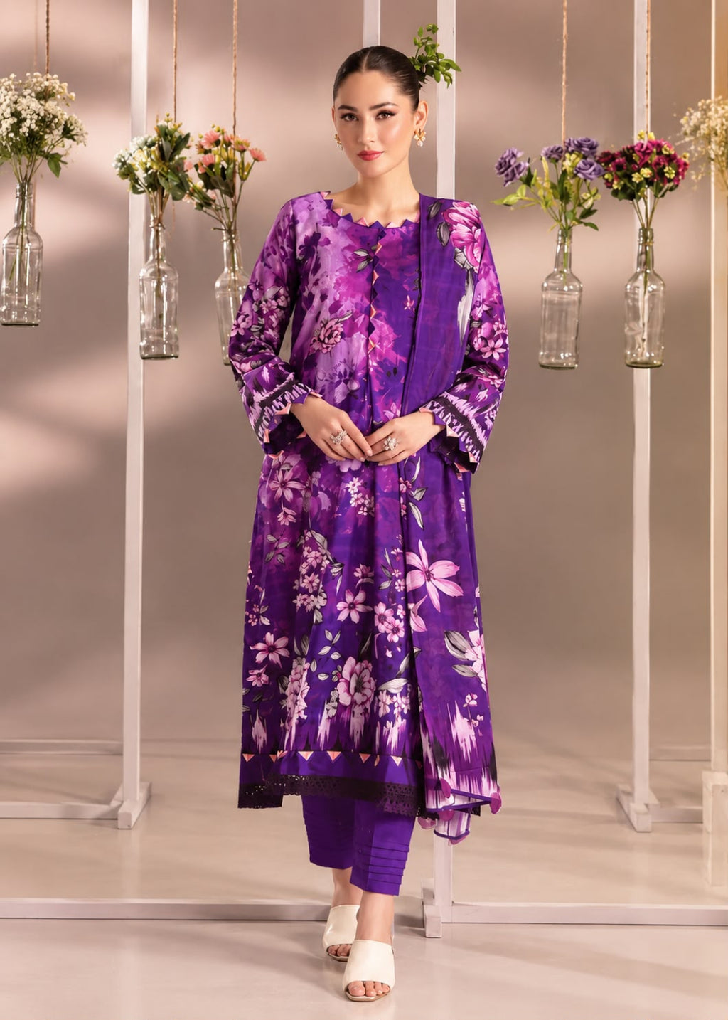A woman models a purple floral printed traditional kurta set with matching trousers, standing in front of a studio backdrop decorated with hanging glass vases of flowers.