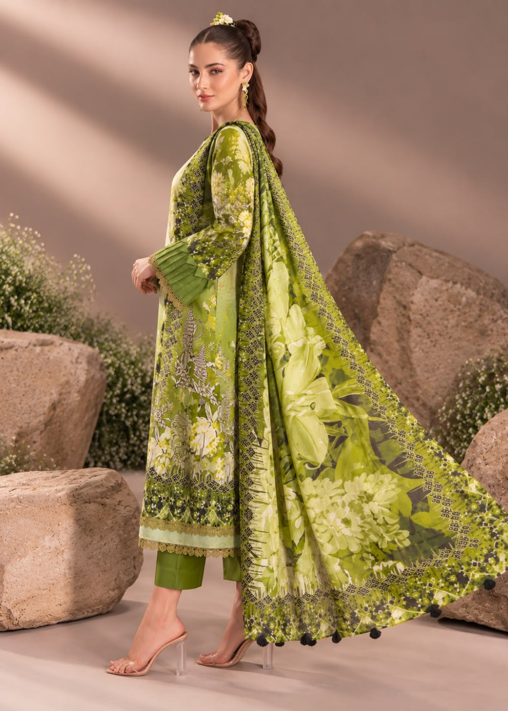 A woman models a three-piece olive green ethnic suit with floral embroidery and a matching dupatta, set against a backdrop of large rocks and plants.