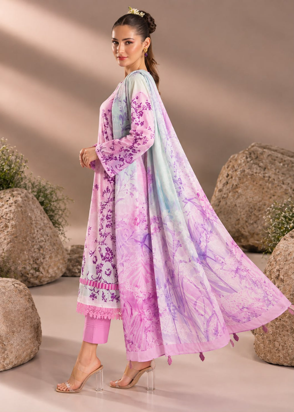A woman models a traditional three-piece salwar kameez suit in a studio setting with decorative rocks. The outfit features a light pink kurta with purple floral embroidery, matching pink trousers, and a sheer light blue and purple printed dupatta.