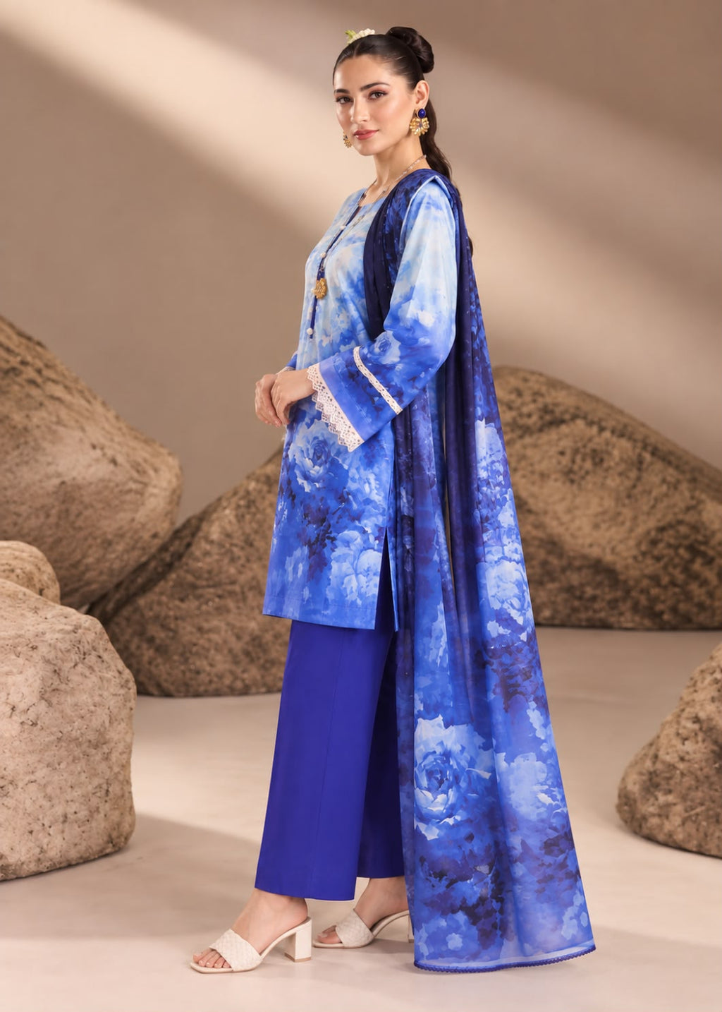 A woman models a blue printed traditional outfit consisting of a long blue tunic with white lace trim, matching royal blue pants, and a long sheer blue overlay, posed in front of a beige backdrop with large decorative rocks.