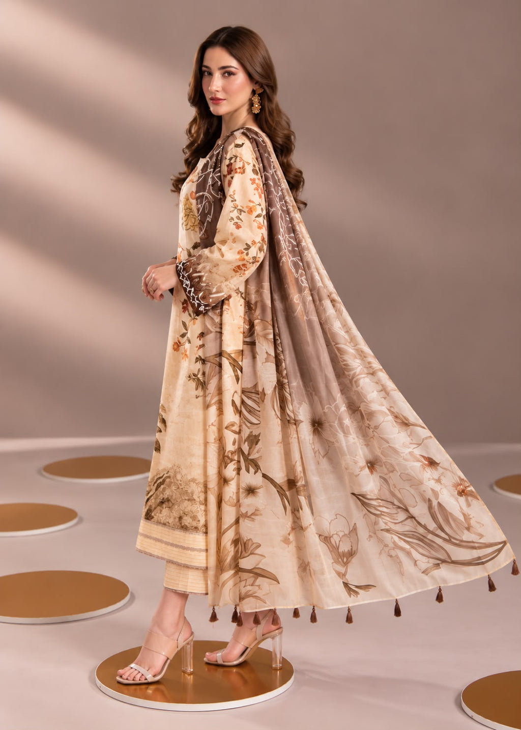 A woman models a traditional beige three-piece outfit with floral prints and a matching dupatta, standing on a circular platform against a neutral studio backdrop.