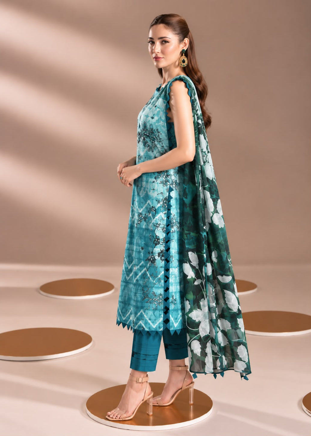 A woman models a teal and green printed salwar kameez suit with a matching printed dupatta, standing on a circular platform against a beige background.