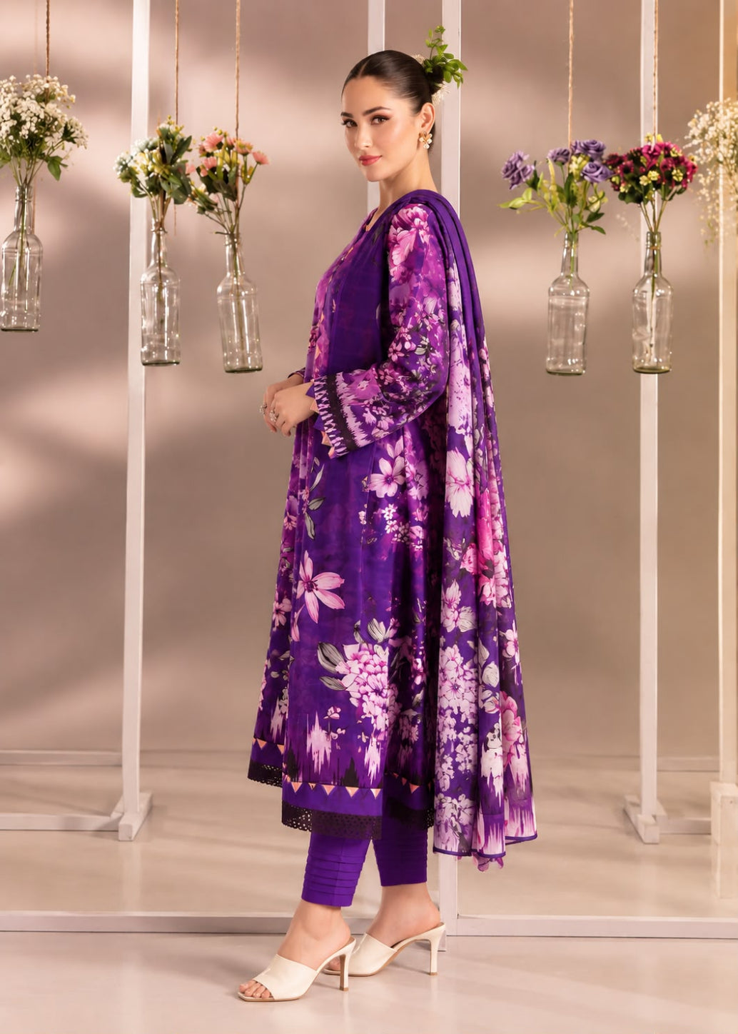 A woman models a traditional South Asian outfit featuring a purple floral kurta with matching trousers and a dupatta, set against a studio backdrop with hanging glass vases.