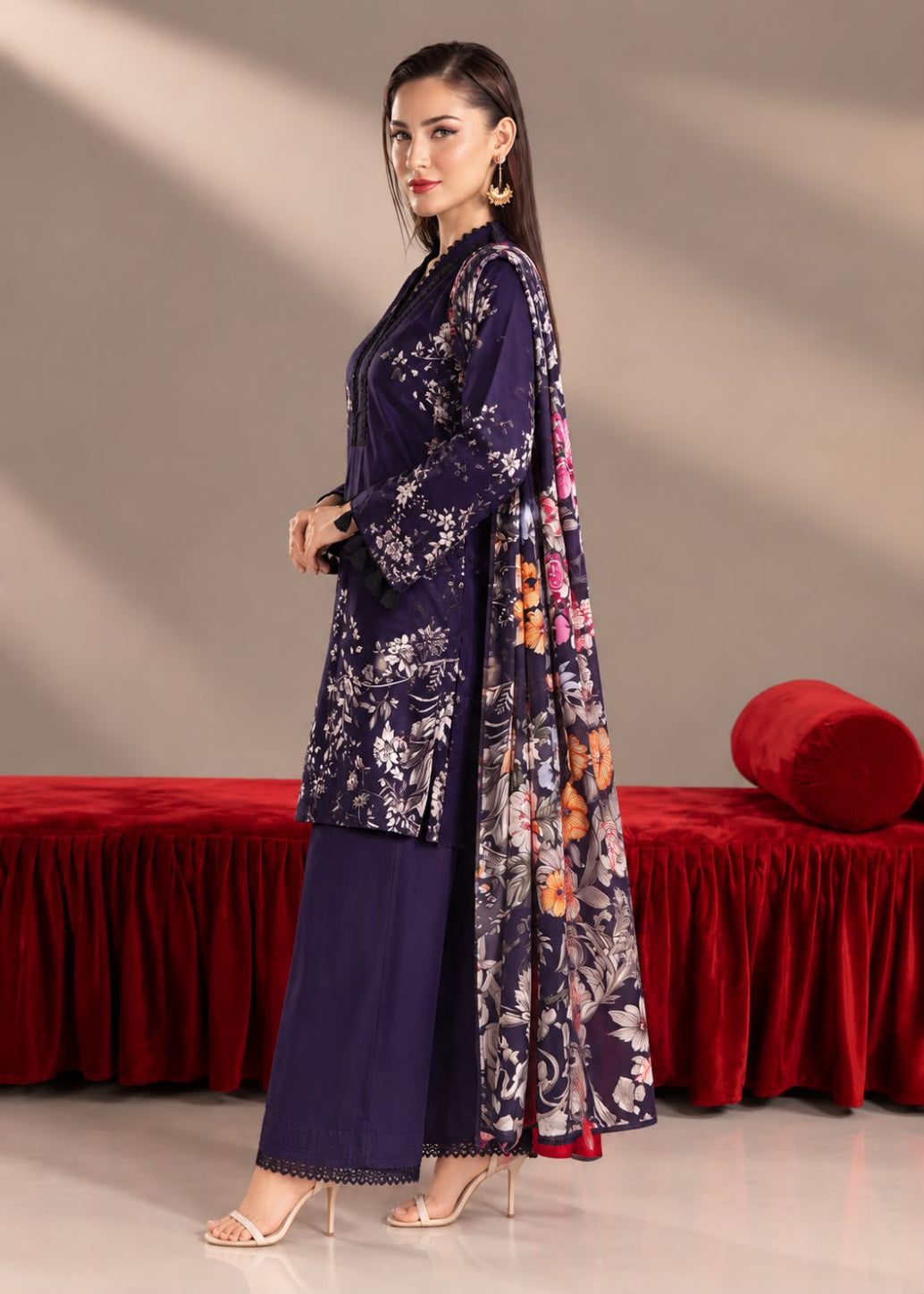 A woman models a traditional South Asian outfit consisting of a deep purple kurta with white floral embroidery, matching wide-leg trousers, and a coordinating long dupatta draped over her shoulder. The background features a beige wall with artistic diagonal shadows and a red velvet bench.