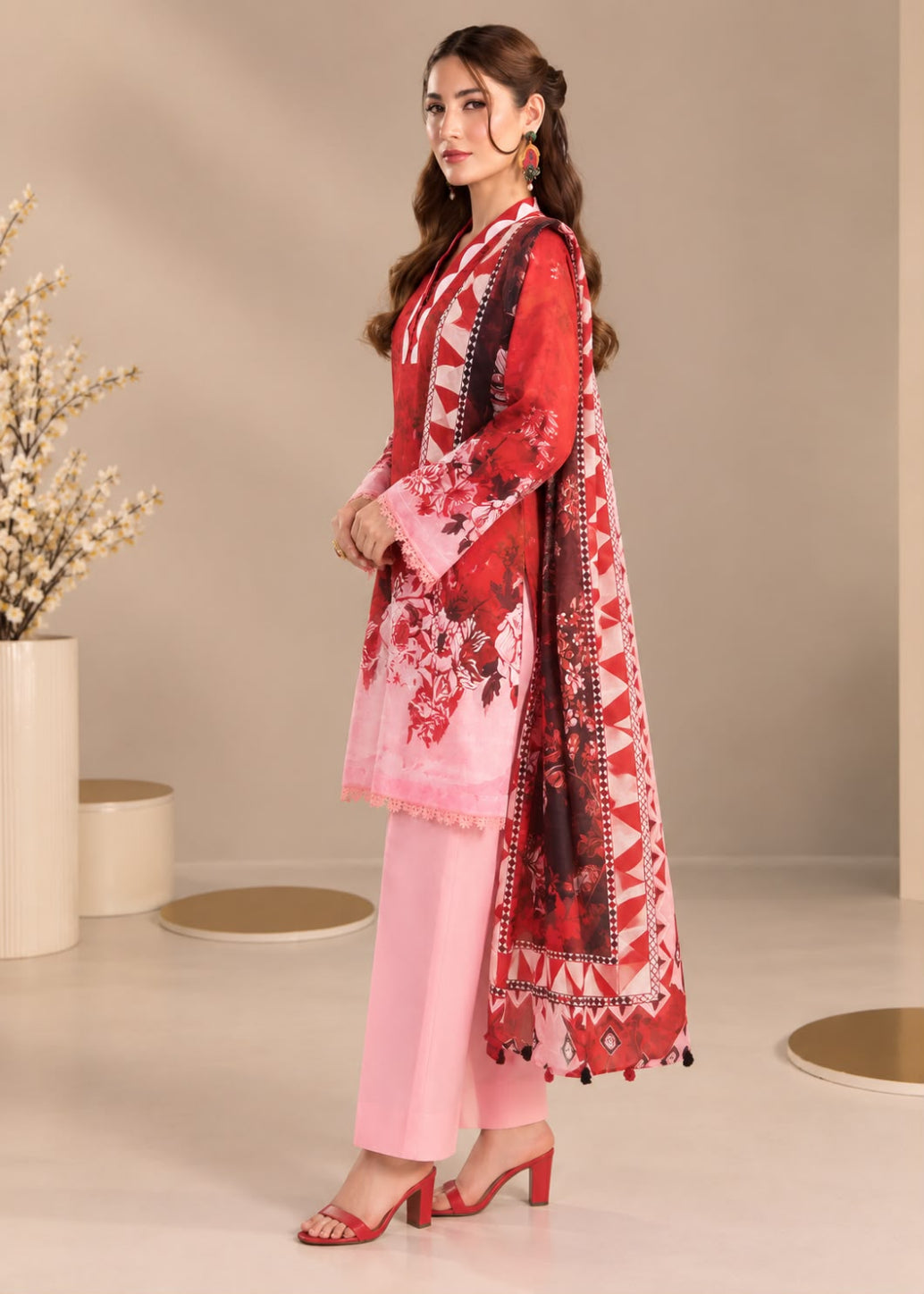 A woman models a three-piece salwar kameez suit set. The set features a light pink kurta with red and black floral patterns and a matching printed dupatta, paired with solid light pink wide-leg trousers and red high-heeled sandals, set against a neutral beige studio background.