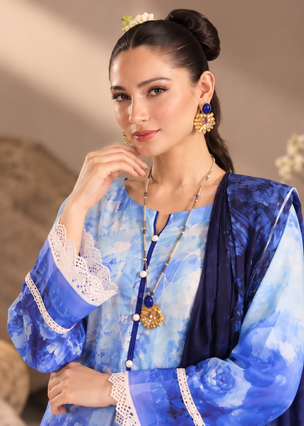 A woman modeling a traditional blue and white printed outfit with a navy blue dupatta, lace-trimmed sleeves, and gold jewelry against a soft beige background.