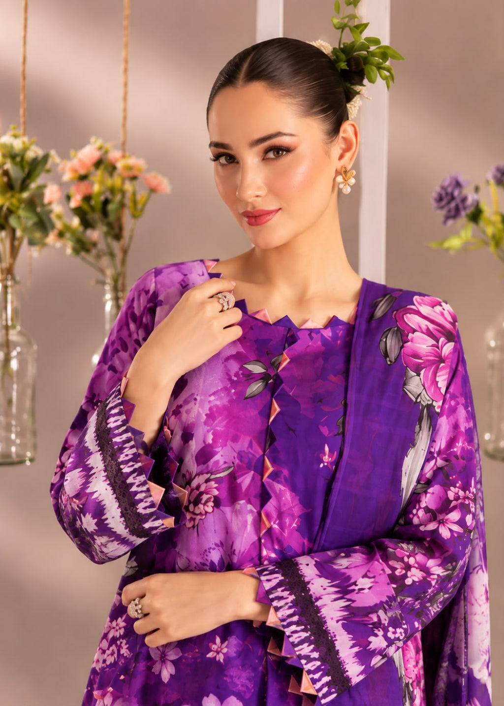 A woman modeling a purple floral printed traditional outfit with long sleeves and a scalloped neckline, posed against a softly lit indoor backdrop with floral arrangements.