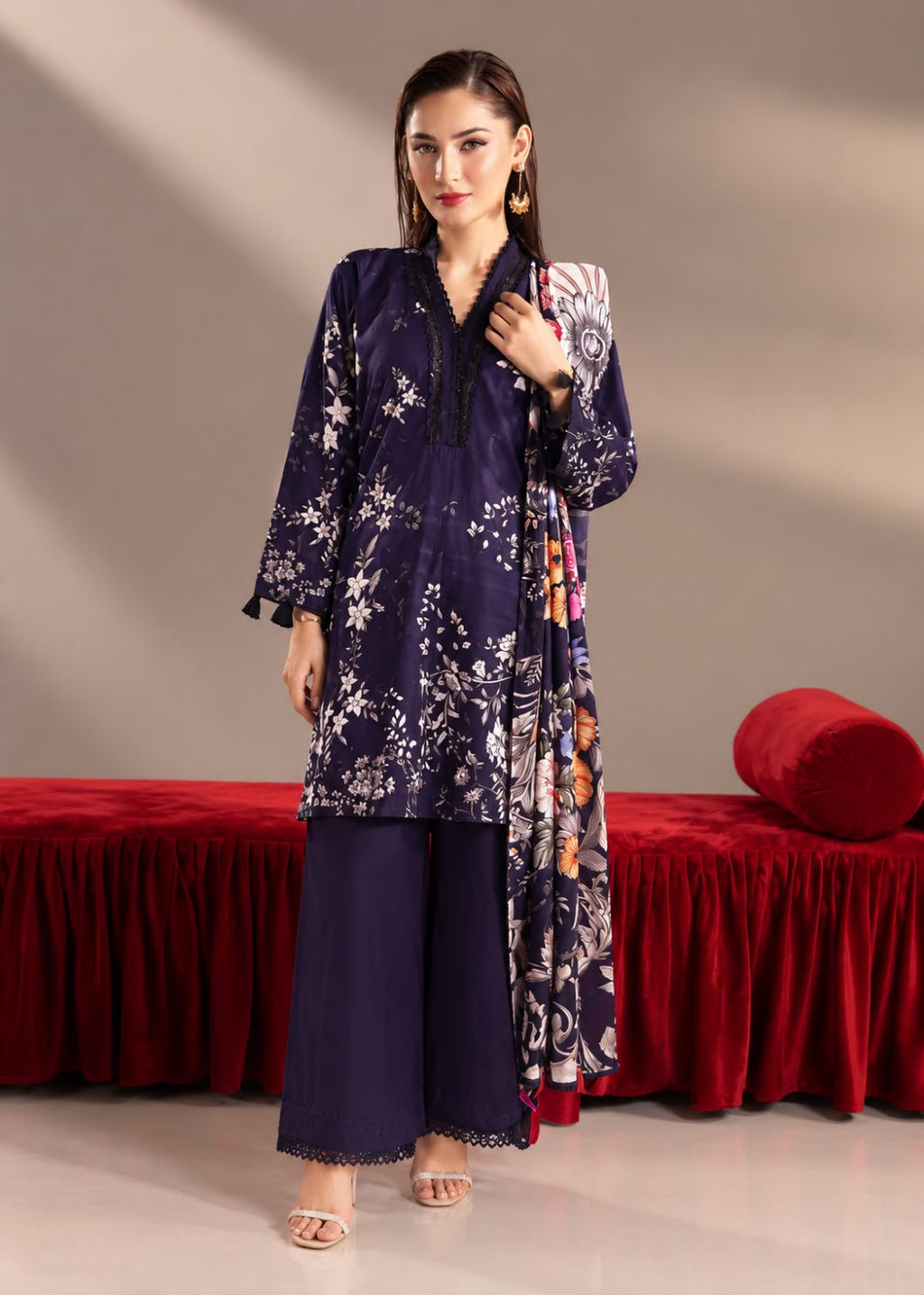 A woman models a three-piece navy blue ethnic salwar kameez suit with white floral prints and a matching printed dupatta, set against a studio backdrop with a red velvet bench.