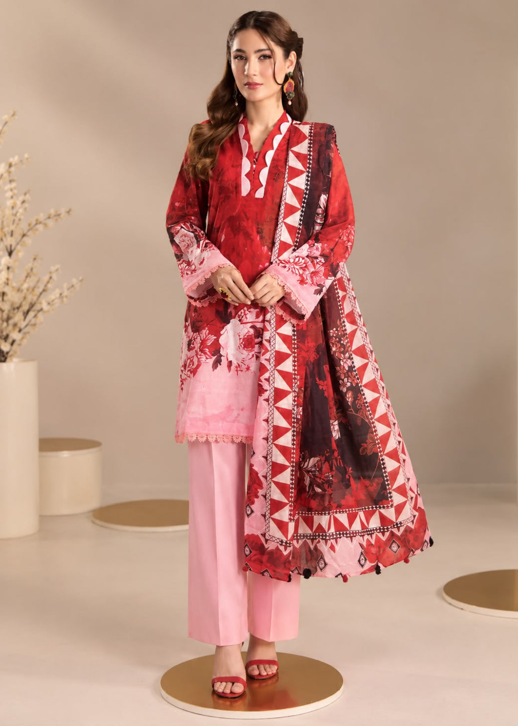 A woman models a three-piece traditional South Asian outfit consisting of a red and pink printed kurta, light pink trousers, and a matching red and pink printed dupatta with geometric borders, set against a neutral beige studio background.