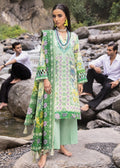 A woman stands barefoot on rocks beside a stream wearing a light green traditional outfit with floral embroidery in yellow, purple, and green. Two men sit on large boulders in the background near the water.