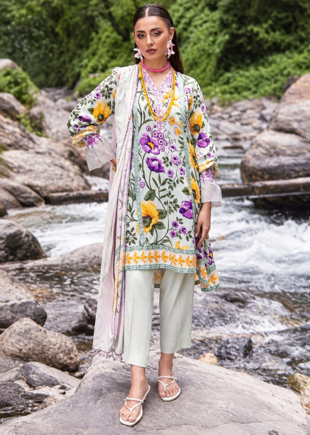 A woman stands on a large rock in front of a rushing stream and green trees, wearing a light sage green three-piece traditional outfit with colorful floral embroidery in yellow, purple, and orange. The set includes a tunic, matching pants, and a draped scarf.