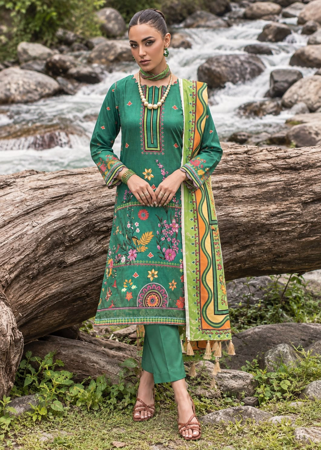 A woman stands outdoors in front of a rocky stream and large log, wearing a green traditional outfit with colorful floral embroidery and a matching patterned dupatta.
