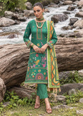 A woman stands outdoors in front of a rocky stream and large log, wearing a green traditional outfit with colorful floral embroidery and a matching patterned dupatta.