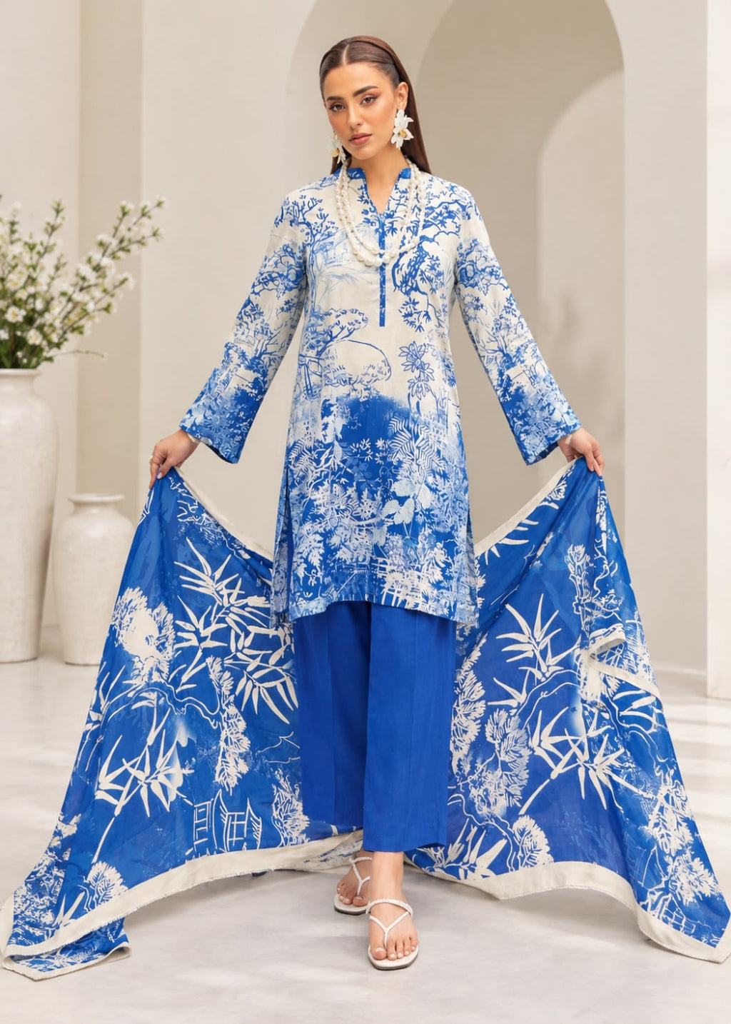 A woman models a three-piece blue and white printed ethnic suit set, featuring a long-sleeve kurta, matching pants, and a dupatta with white bamboo and floral prints, set against a neutral indoor background.