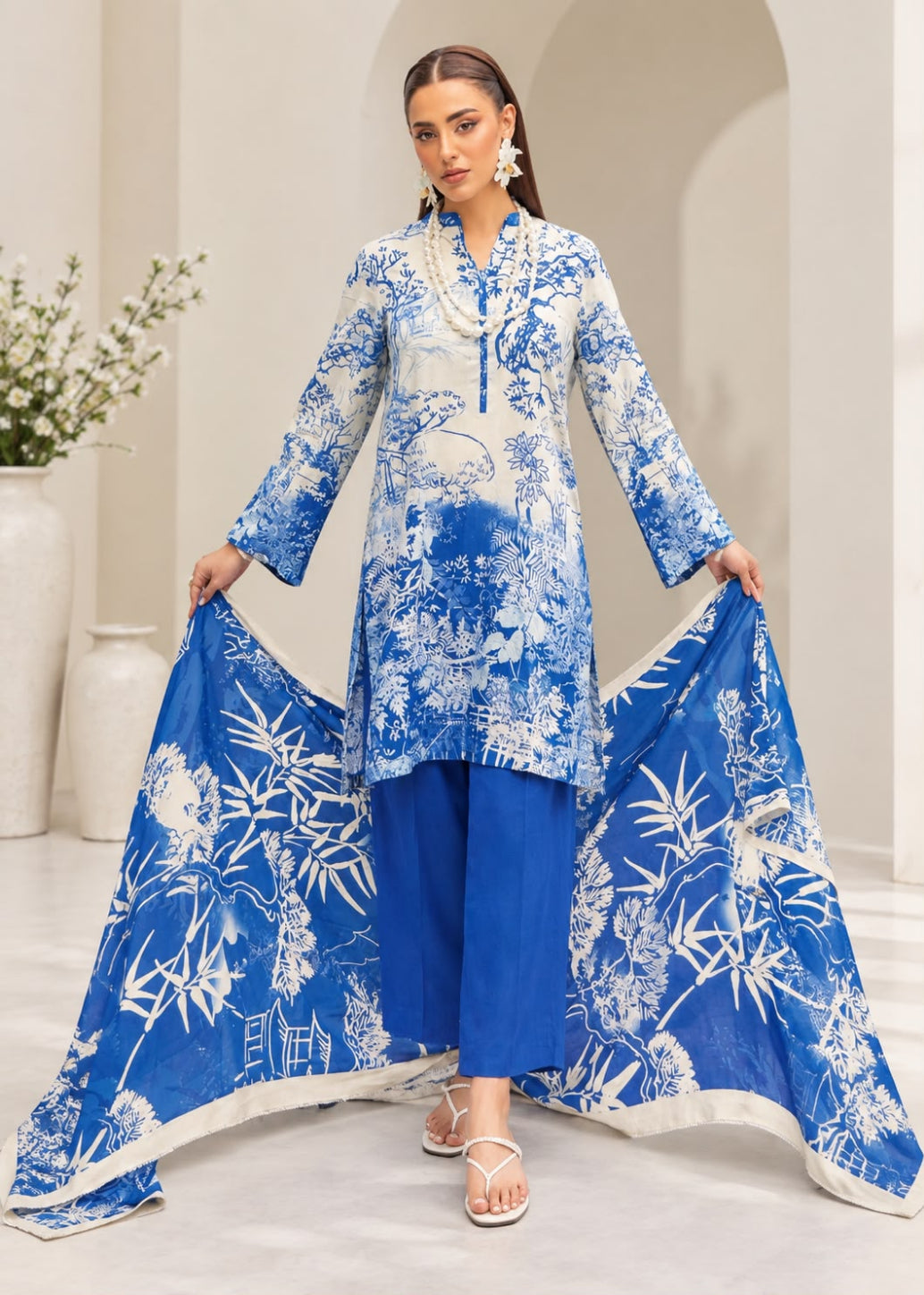 A woman models a three-piece blue and white printed ethnic suit set, featuring a long-sleeve kurta, matching pants, and a dupatta with white bamboo and floral prints, set against a neutral indoor background.