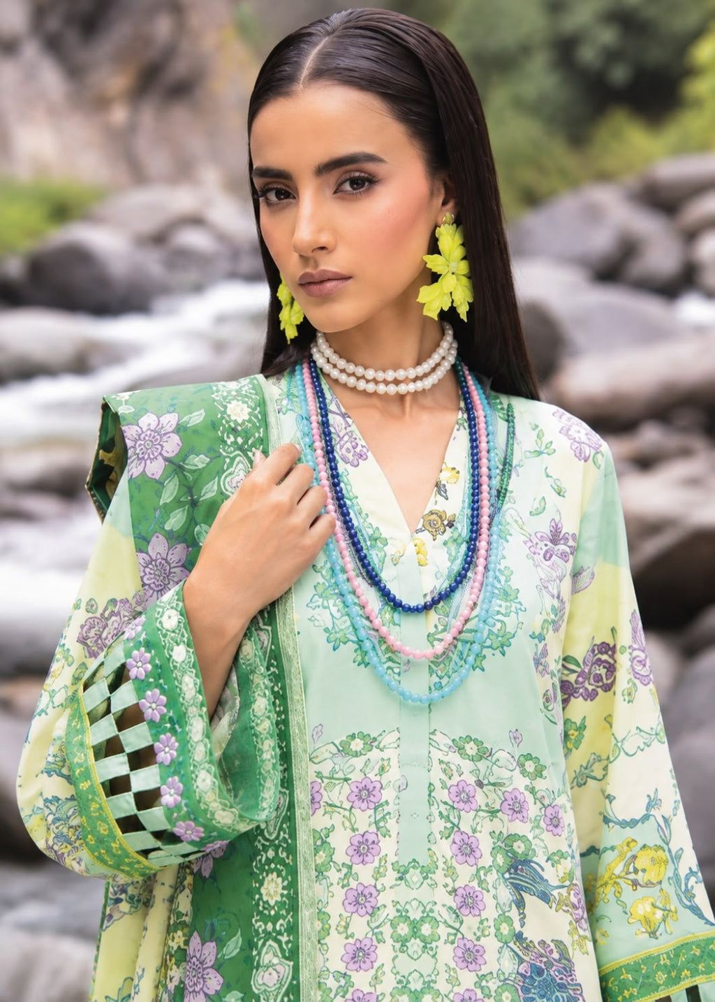 A woman wearing a light green and white printed traditional outfit with floral patterns, layered beaded necklaces, and bright yellow earrings, posed outdoors in front of a blurred rocky stream background.