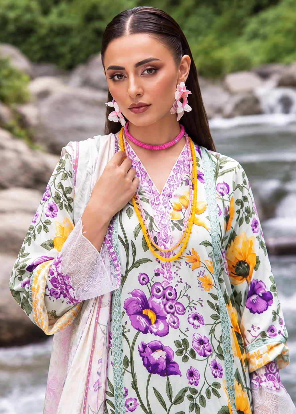 A woman wearing a floral print traditional outfit with purple and yellow flowers, green leaves, and pink accents, standing outdoors in front of a blurred rocky stream background.