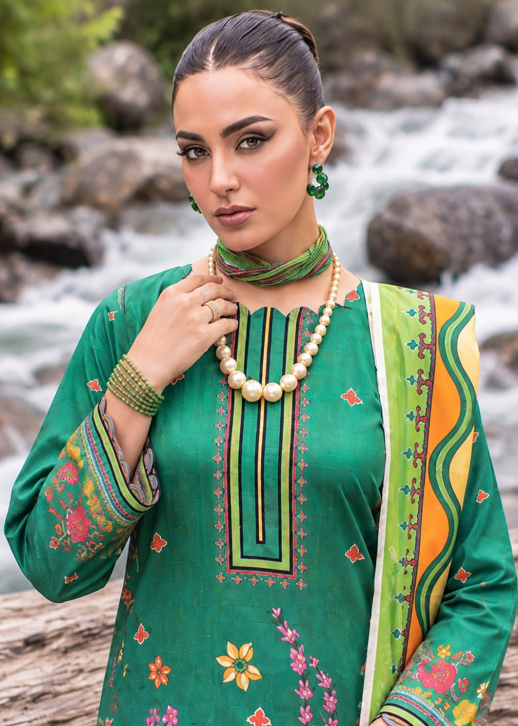 A woman wearing a green embroidered traditional outfit with a matching dupatta, standing outdoors in front of a flowing river with rocks in the background.