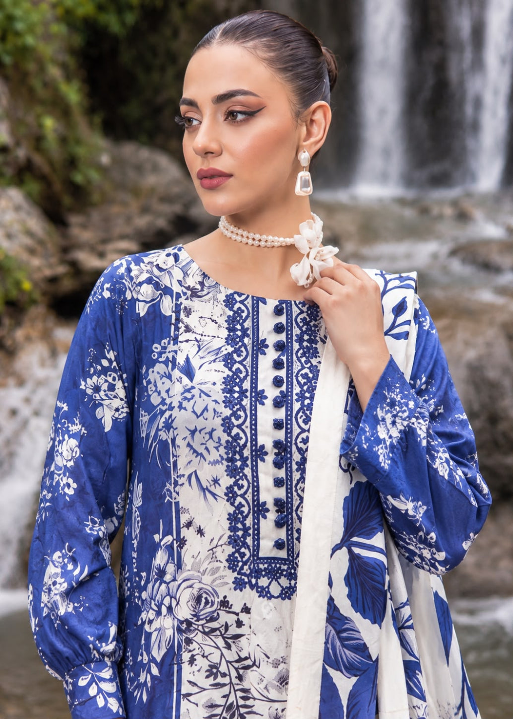 A woman wearing a blue and white printed traditional outfit with a matching dupatta, standing outdoors in front of a waterfall and rocks.