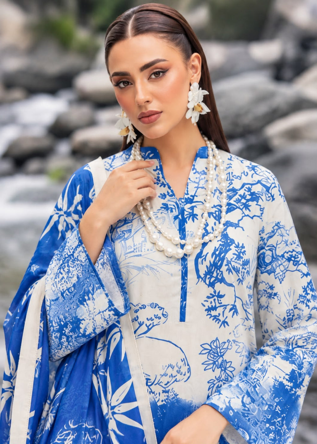 A woman wearing a blue and white printed traditional outfit with a matching dupatta, layered white pearl necklaces, and white flower earrings, posed outdoors against a blurred rocky background.