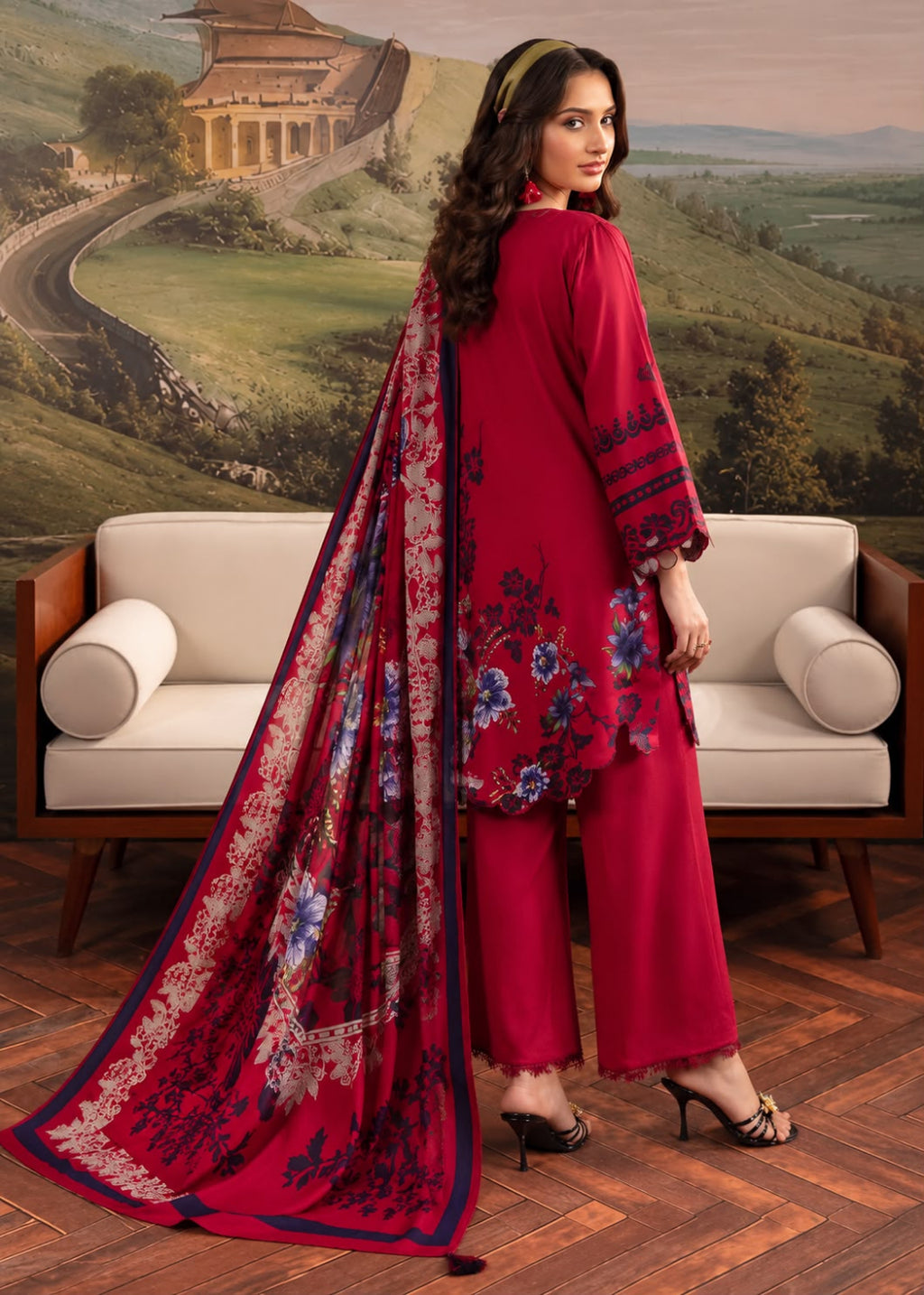 A woman models a three-piece traditional Indian salwar kameez suit in a vibrant red color, featuring a floral printed dupatta and matching printed trousers. She is posed in front of a white sofa with a scenic landscape mural in the background.