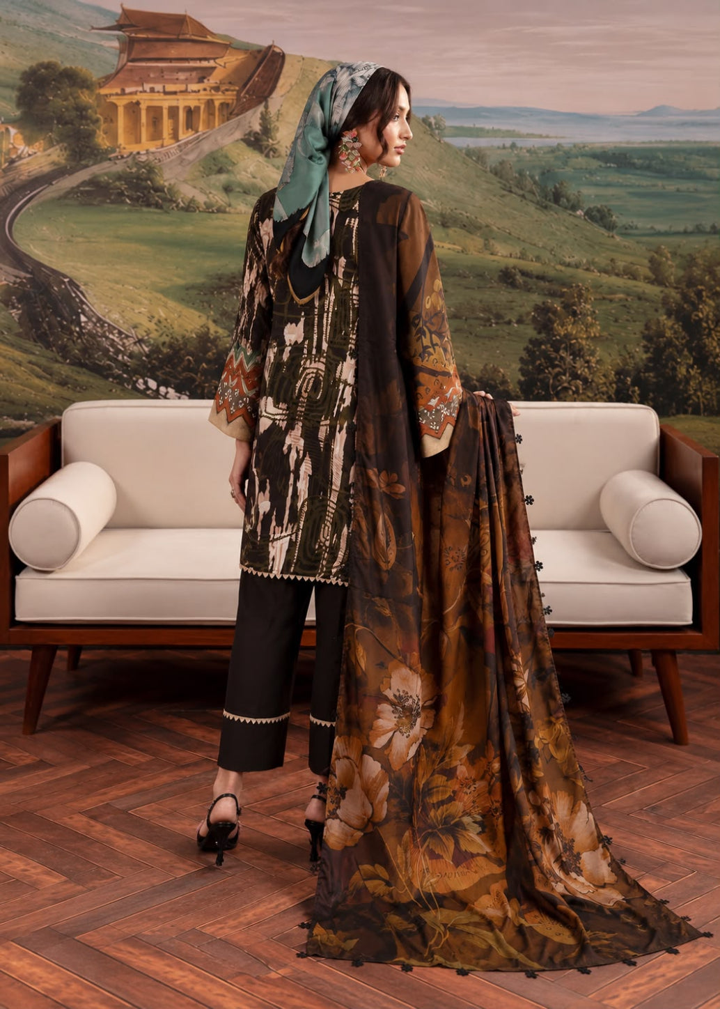 A woman models a traditional South Asian outfit consisting of a brown and black patterned kurta, black trousers, and a matching brown floral dupatta, posed in front of a white sofa with a scenic landscape mural background.