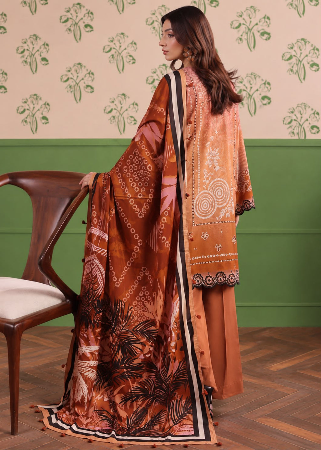 A woman wearing a traditional South Asian outfit with a rust-orange printed dupatta, matching trousers, and a patterned jacket, standing in a room with a green and beige patterned wall and a wooden chair.