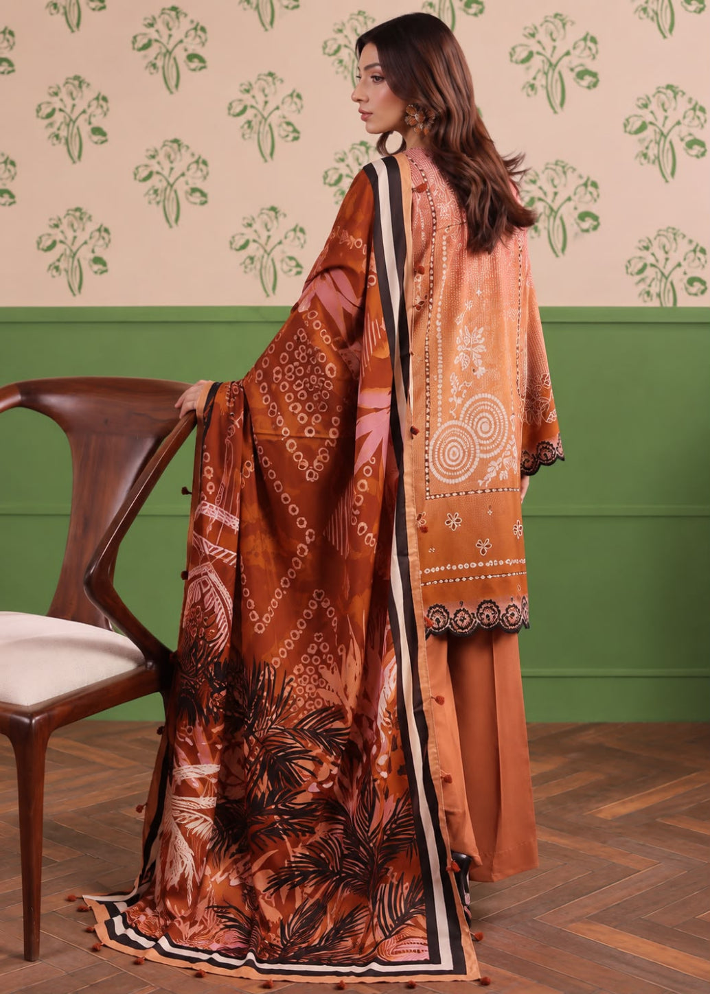 A woman wearing a traditional South Asian outfit with a rust-orange printed dupatta, matching trousers, and a patterned jacket, standing in a room with a green and beige patterned wall and a wooden chair.